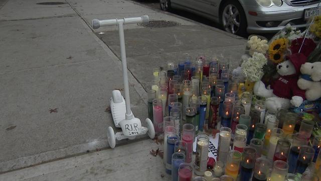 A large collection of candles, stuffed animals, flowers and a small scooter sit on a street corner. 
