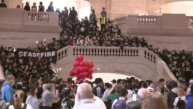 Dozens of demonstrators, many wearing black t-shirts that read "Not in our name" or "Cease fire now," stand on the staircase inside Grand Central Terminal. 
