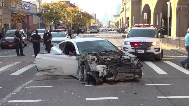 A vehicle with severe front-edge damage sits in a crosswalk while NYPD officers stand in the background. 