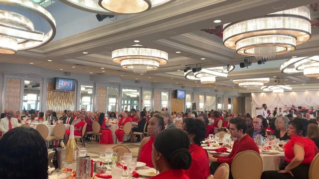 A few hundred people sit at round tables in a banquet hall. Many are wearing red.