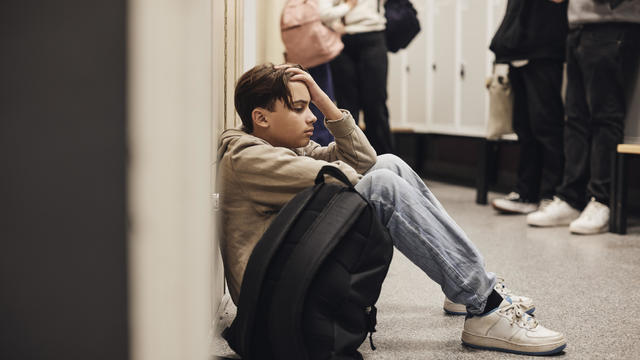 Side view of sad teenage boy sitting with backpack in school corridor 