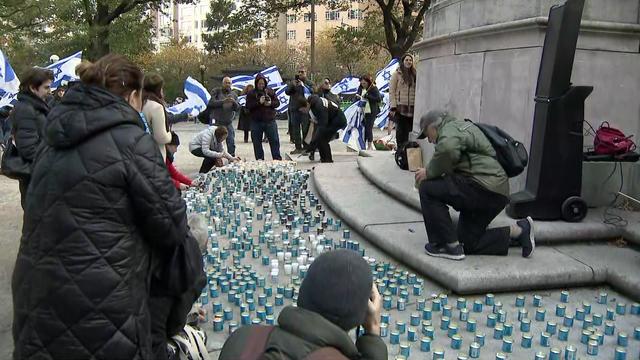 Hundreds of candles sit on the ground at the base of a statue in Columbus Circle. Numerous people holding Israeli flags stand in the background. 