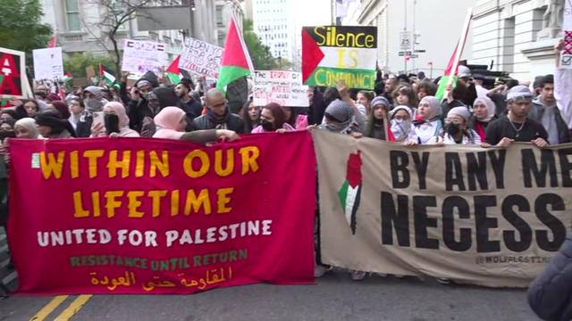 Hundreds of demonstrators stand in a street behind two banners with pro-Palestinian messaging. 