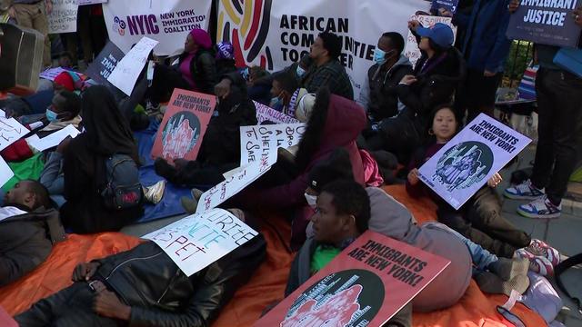 Multiple individuals lay on a sidewalk outside Gracie Mansion, many on sleeping bags or holding signs. 