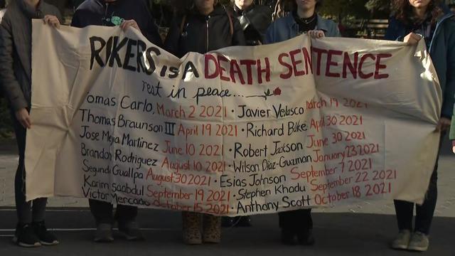 Individuals hold a banner that says "Rikers is a death sentence," followed by the names of individuals who have died on Rikers. 