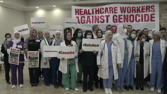 Dozens of medical professionals stand in front of a banner reading, "Healthcare Workers Against Genocide." 