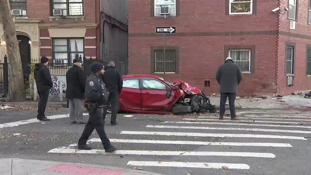 NYPD officers stand around a red sedan with severe front-end damage on a Bronx street. 