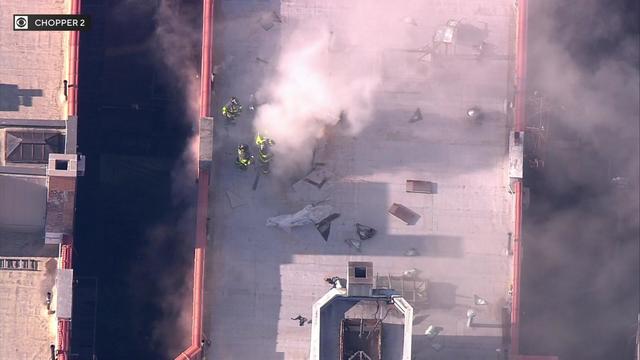 Firefighters work on the roof of an apartment building as smoke pours out a hole in the roof. 