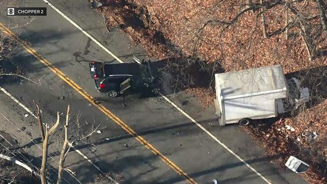 An aerial view of a black SUV with front-end damage sitting in the middle of the road. Nearby, a white box truck with front end damage sits off the side of the road. 
