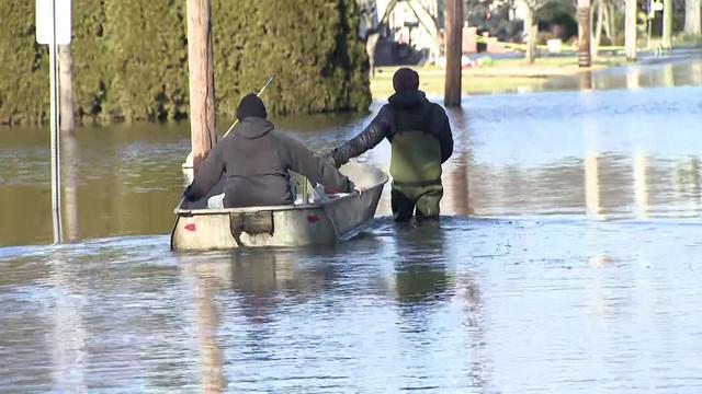 A man sits in an aluminum dinghy as another man pulls it through floodwaters. 