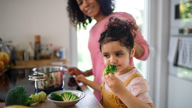 Little girl eating preparing broccoli for dinner with her mother in kitchen. 