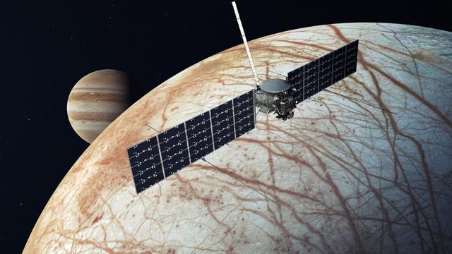 Illustration showing the Europa Clipper, a spacecraft with two solar panels, flying by Europa with Jupiter in the background