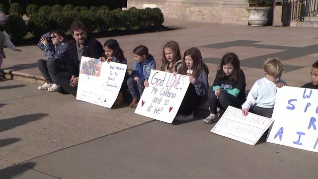 Michael Califano sits on a curb with several children. Many children hold signs reading "God LOVES Mr. Califano and so do we!" and "BRNG BACK MR. CALIFANO." 