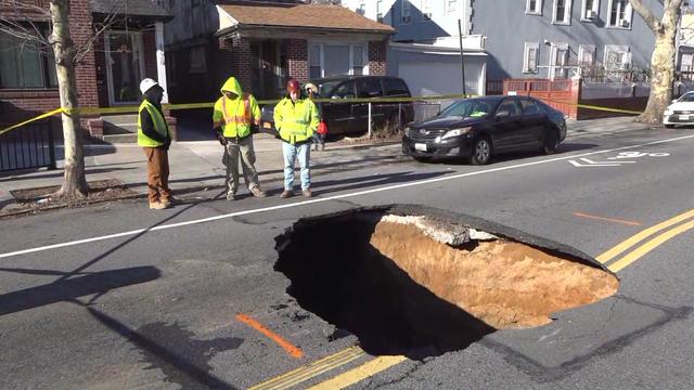 Three workers in reflective vests stand at the edge of a sinkhole in the middle of a road in Brooklyn. 