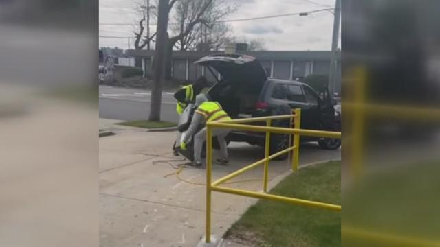 Two individuals wearing construction vests lift an ATM into the back of an SUV. 