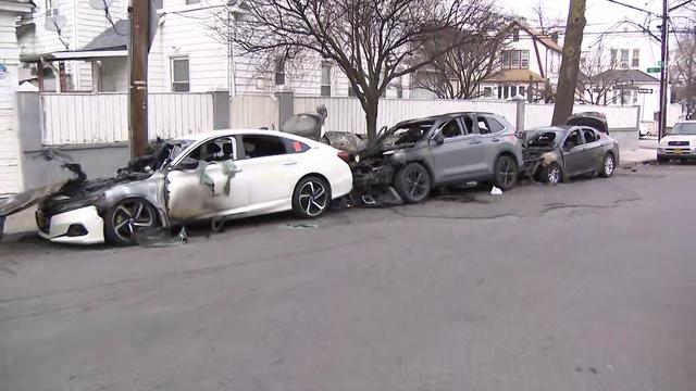 Three vehicles parked in a row on a street in Queens. All three vehicles have fire damage. 