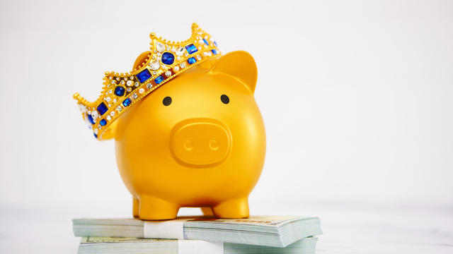 Gold piggy bank wearing crown standing on a stack of American currency 