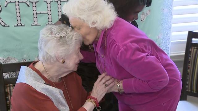 A woman kisses Lottie Higgins on the forehead. Lottie sits in a chair wearing a plastic tiara and a sash. 