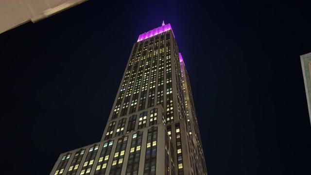 A view of the exterior of the Empire State Building from street level. The top of the building is lit up in bright purple. 