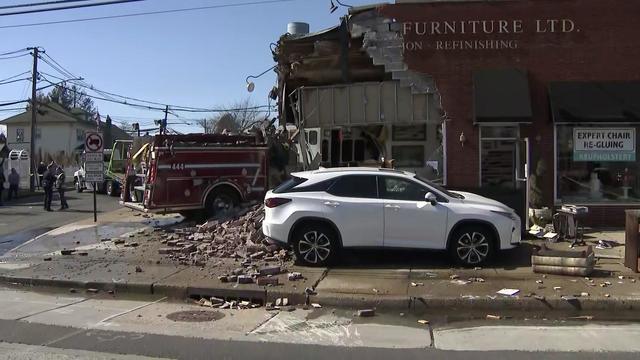 A large portion of the corner of a brick building has been destroyed. A fire truck sits partially inside the building. A white SUV sits in front of the building. 