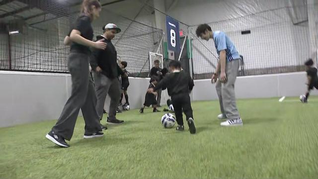 Talya Landesberg, Power Malu and a teenager follow a small child kicking a soccer ball on an indoor field. 
