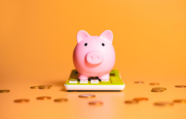 A piggy bank over a green calculator with scattered coins isolated on orange background in studio photography. Concepts of saving money, finance and investments. 