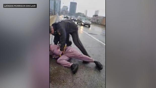A screenshot of cellphone video showing a person being handcuffed on the side of a highway 