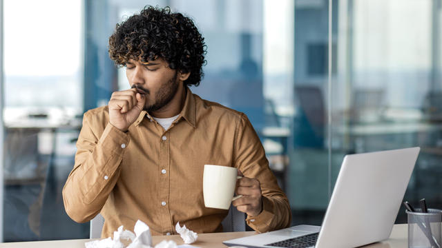 Sick businessman at workplace, man coughing and sneezing, cold and runny nose, sitting at desk inside office, holding cup of hot drink. 