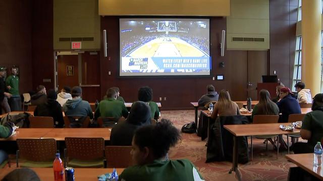 Individuals sit at tables watching a basketball game on a screen. 