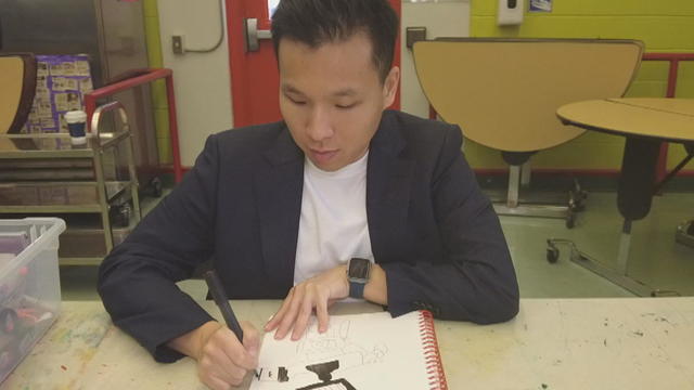 Suwon Cho is sitting at a table as he draws in a notebook 