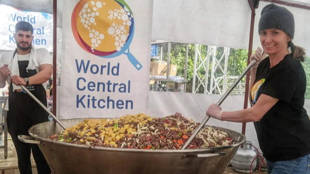 Two people hold huge spoons, stirring a giant bowl in front of a sign that says World Central Kitchem 