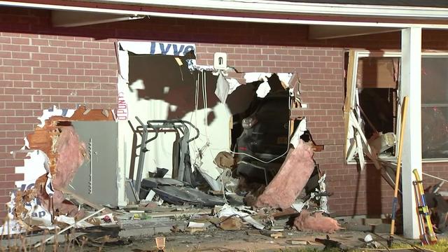 An SUV sits completely inside a home, visible through a large hole in a brick wall. 