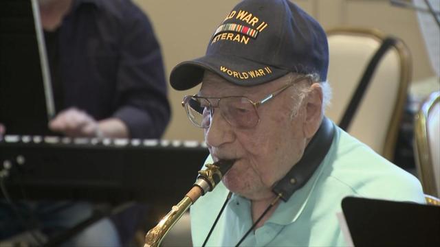 Dominick Critelli sits with an orchestra in a practice room, playing saxophone and wearing a World War II veteran baseball hat. 
