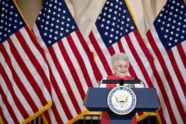 Mae Krier speaks at a podium in front of several American flags 