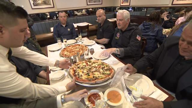 PBA President Thomas Shevlin, Nassau County Executive Bruce Blakeman and others sit around a table as a waiter sets a pizza down on the table. 