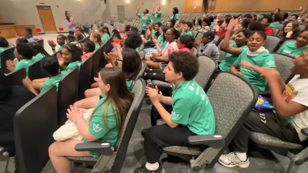 A group of middle schoolers in a school auditorium clap during a presentation. They're all wearing green shirts 