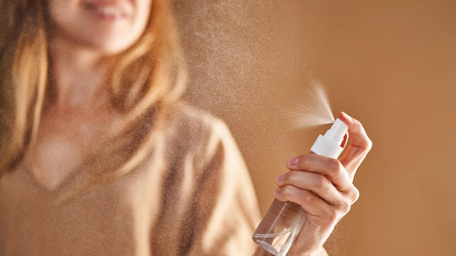A girl in a beige dress sprays tonic or thermal water from a transparent bottle. 