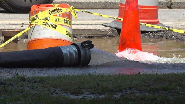 Water pours out of a large hose into a street near several orange cones and caution tape. 