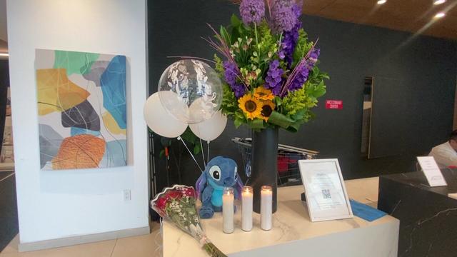 Bouquets of flowers, balloons, candles and a stuffed animal Stitch sit on a countertop in the lobby of a Queens apartment building. 