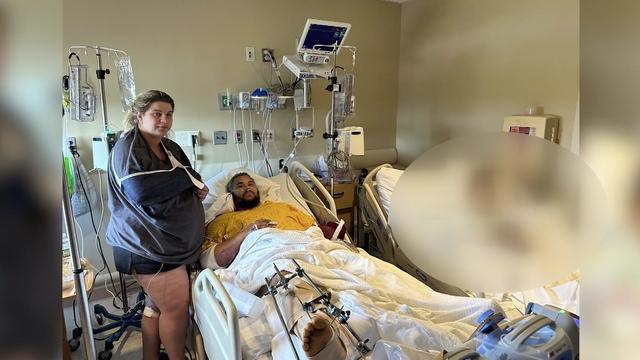 Ashley Ambroise stands next to her husband, who is laying in a hospital bed. Ashley's right arm is in a sling and she is nine months pregnant. Her husband's right leg is injured. 