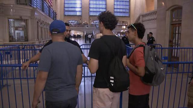 Inside Grand Central Terminal, an MTA Police officer speaks to three members of the public. The police officer and individuals are separated by a barricade. Multiple other barricades are visible behind the officer. 