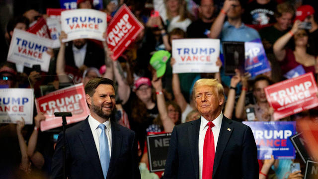 JD Vance and Donald Trump stand on stage at a rally, supporters hold up Trump-Vance signs behind them 