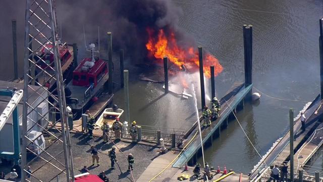 Firefighters spray water on a boat engulfed in flames in the water at a marina. 