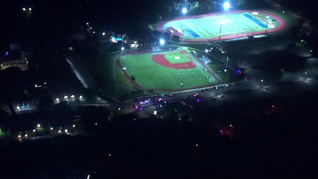 An aerial view of a parking lot next to a baseball field in Wayne, New Jersey. Crime scene tape blocks off the lot and evidence markers can be seen on the sidewalk. 