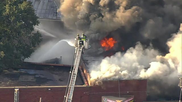 Flames and smoke pour from the roof of a building in Queens as firefighters on a ladder spray water. 