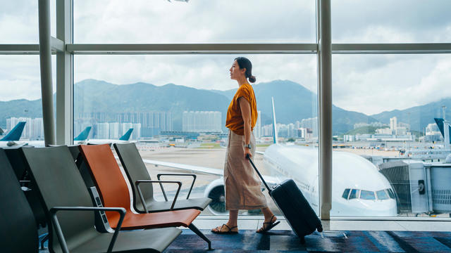 Young Asian woman carrying suitcase, walking by the window at airport terminal. Young Asian female traveller waiting for boarding at airport. Business travel. Travel and vacation concept 