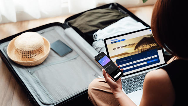 Over the shoulder view of young woman booking a flight via laptop and smartphone for vacation 