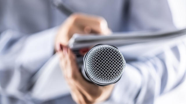 A journalist holds a microphone at a press conference and writes information in a notebook. 