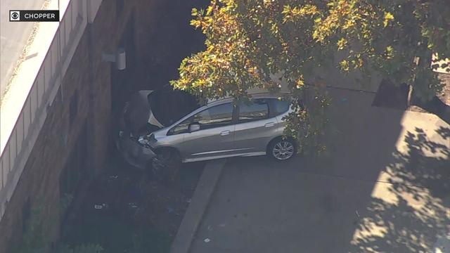 An aerial view of a silver four-door sedan that has crashed into the side of a school, causing severe front-end damage to the vehicle. 