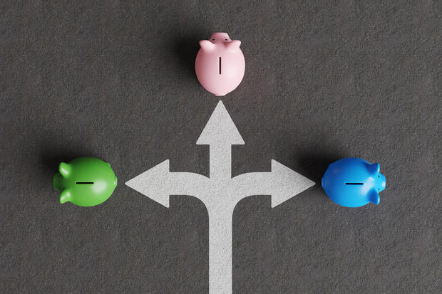 White divided road sign mark on asphalt with 3 different colored piggy banks going to different directions. Illustration of the concept of stock picks and varied investment products 